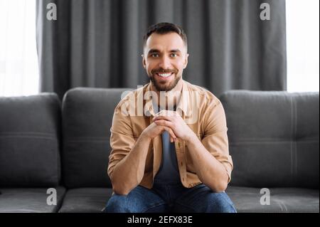 Ritratto di un attraente ragazzo caucasico vestito in elegante abbigliamento casual. Uomo sicuro guarda direttamente la macchina fotografica con un sorriso amichevole mentre si siede sul divano in un soggiorno Foto Stock