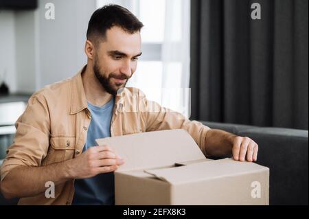 Consegna a domicilio. Bel ragazzo caucasico stile sedersi sul divano, disimballare scatola di cartone, sorriso. L'uomo soddisfatto ha ottenuto un pacchetto atteso lungo, era shopping nei depositi del Internet, concetto di shopping in linea Foto Stock
