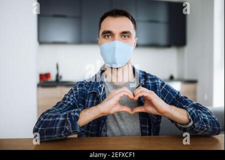 Misure preventive. Un ragazzo caucasico in una maschera medica protettiva si siede a casa in un abbigliamento casual, fa un gesto del cuore con le mani e guarda direttamente nella fotocamera Foto Stock