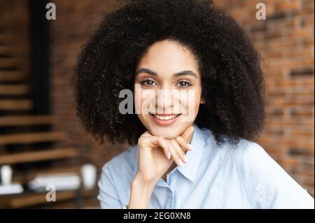 Ritratto headshot di giovane afroamericana intelligente etnia femminile con afro curly acconciatura e bel sorriso, messo mano sul mento, guardando Foto Stock