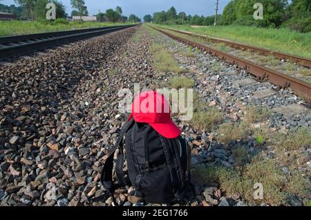 Uno zaino nero con due cinturini e berretto rosso si trova tra due binari ferroviari con traversine su uno sfondo di prato, alberi e blu chiaro Foto Stock