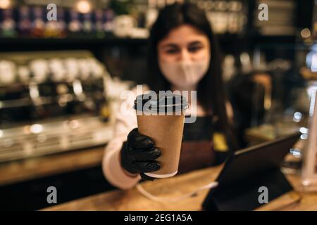 Il barista in maschera protettiva e guanti neri offre una tazza di caffè al bar. Foto di alta qualità Foto Stock