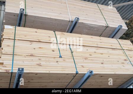 Impaccate in file e pile di travi di legno legate su supporti orizzontali zincati, immagazzinate in un magazzino sotto tetto in metallo corrugato. Foto Stock