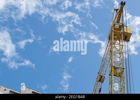 Una gru a torre gialla lavora in un cantiere su i piani superiori di un edificio in mattoni bianchi contro un cielo blu con nuvole fumose e sguazzate Foto Stock