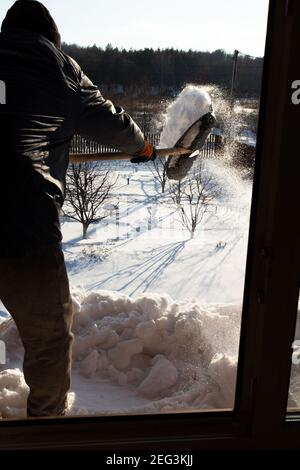 Un uomo pulisce la neve dal tetto di una casa di campagna. Uno sguardo attraverso la finestra del giardino innevato. Shot verticale Foto Stock