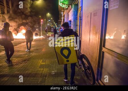 Barcellona, Catalogna, Spagna. 17 Feb 2021. L'addetto alla consegna di cibo a casa dell'azienda, Glovo, è visto di fronte a container in fiamme.seconda notte di manifestazioni, a Barcellona, per la prigione del rapper catalano, Pablo Hasél, arrestato martedì, Febbraio 16 e condannato a nove mesi e un giorno di prigione dalla Camera d'appello della Corte nazionale nel settembre 2018, nonché il pagamento di un'ammenda di circa 30,000 euro accusata di glorificazione del terrorismo, insultare e calunniare la monarchia e le forze di sicurezza dello stato. (Credit Image: © Thiago Prudencio/DAX via ZUMA Wir Foto Stock