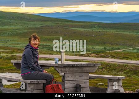Donna seduta su una panchina sul Monte Dundret ha bevuta una tazza di caffè al tramonto, Gällivare, Lapponia svedese, Svezia