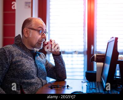 Un uomo caucasico di mezza età che lavora da casa, mentre beve un caffè, è piacevolmente sorpreso da una notifica che legge sul suo monitor del computer. Foto Stock