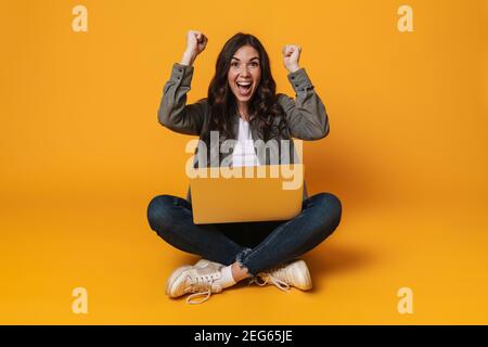 Ragazza entusiasta che fa il gesto del vincitore e che usa il laptop mentre si siede su pavimento isolato su sfondo giallo Foto Stock