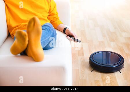 Uomo che riposa mentre l'aspirapolvere robot fa i compiti, lavoro pulito a casa. Man controlla un aspirapolvere robot utilizzando un telecomando. Robot pulitore v Foto Stock