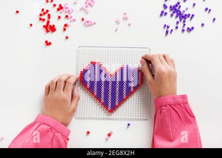 Le mani del bambino piccolo fanno l'arte delle perle nella forma del cuore. Concetto di educazione dei bambini. Artigianato per bambini. Vista dall'alto. Foto Stock