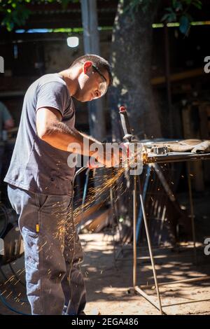 un uomo che lavora tagliando una barra metallica con un elettrico circolare strumento che spara scintille Foto Stock