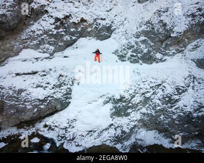 Drone colpo di escursionista con zaino, cappello arancione e pantaloni arancio trekking su pericoloso sentiero attraverso la neve e ghiaccio vicino al freddo fiume di montagna Foto Stock