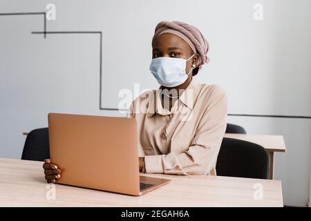 Donna d'affari musulmana nera in maschera medica con computer portatile. Lavorare in linea a casa. Investimenti di criptovaluta in bitcoin. Ragazza nera riuscita. Foto Stock