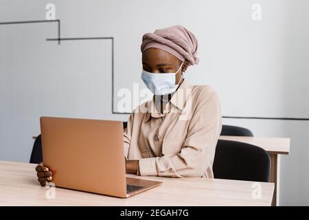 Donna d'affari musulmana nera in maschera medica con computer portatile. Lavorare in linea a casa. Investimenti di criptovaluta in bitcoin. Ragazza nera riuscita. Foto Stock