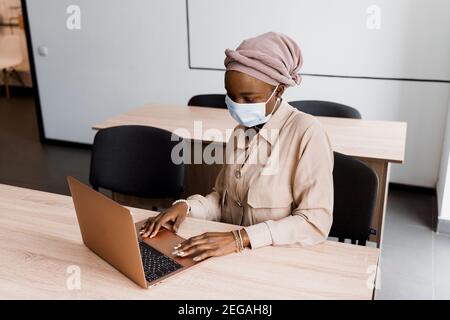 Donna d'affari musulmana nera in maschera medica con computer portatile. Lavorare in linea a casa. Investimenti di criptovaluta in bitcoin. Ragazza nera riuscita. Foto Stock