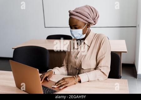 Donna d'affari musulmana nera in maschera medica con computer portatile. Lavorare in linea a casa. Investimenti di criptovaluta in bitcoin. Ragazza nera riuscita. Foto Stock