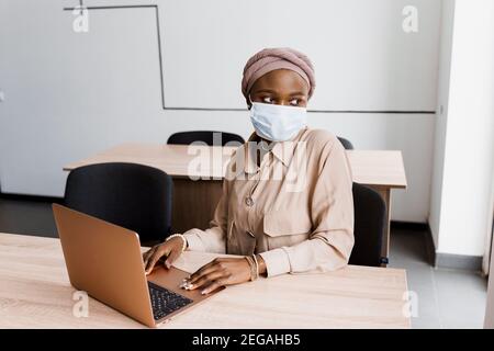 Donna d'affari musulmana nera in maschera medica con computer portatile. Lavorare in linea a casa. Investimenti di criptovaluta in bitcoin. Ragazza nera riuscita. Foto Stock