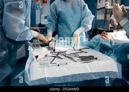 primo piano chirurghi che prendono a mano forbici, pinzette e strumenti chirurgici sul tavolo per l'uso con i colleghi che eseguono il lavoro in sala operatoria alle ore Foto Stock