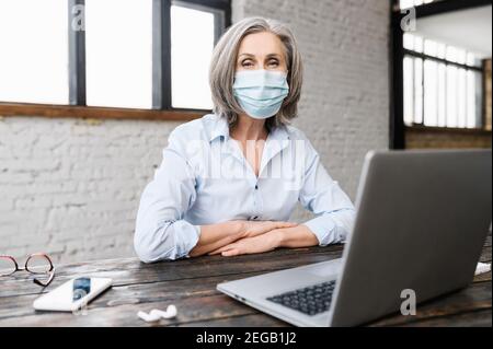 Donna anziana sicura che indossa la maschera sul posto di lavoro in ufficio. Anziana dipendente femminile si protegge durante la pandemia di covidio seduta a. Foto Stock