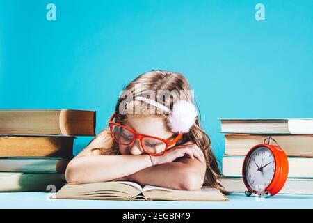 Bambina che dorme su un libro aperto in occhiali rossi divertenti con pile di libri accanto a lei. Ritorno a scuola. Foto Stock