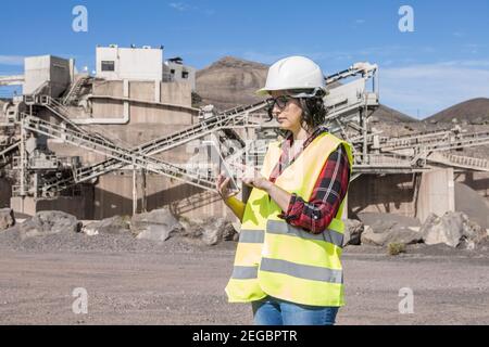 Vista laterale di una donna specializzata in elmetto e gilet controllo delle informazioni sul tablet mentre ci si trova vicino a una struttura industriale di costruzione s Foto Stock