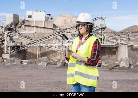 Architetto femminile professionista in elmetto e gilet che controlla la bozza tablet in piedi in prossimità di impianti industriali di cantiere Foto Stock