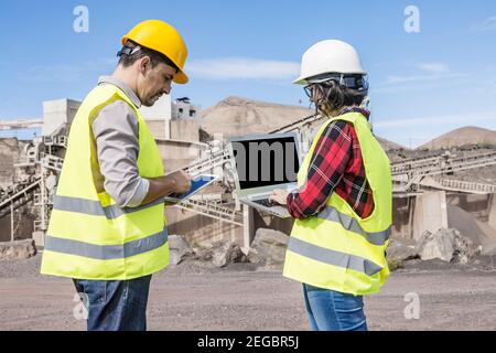 Vista laterale di un ingegnere maschile focalizzato che utilizza tablet e donne collega che scrive sul computer portatile mentre si sta in piedi insieme contro una struttura industriale Foto Stock