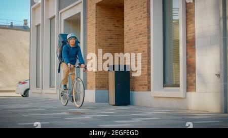 Happy Food Delivery uomo che indossa lo zaino termico su una bicicletta consegna pizza ordine a un cliente. Il corriere consegna il pranzo al Takeaway al moderno quartiere della città Foto Stock