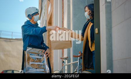 Food Delivery l'uomo che indossa una maschera medica protettiva e uno zaino termico su una bicicletta consegna l'ordine a un cliente Masked Female. Corriere consegna Foto Stock
