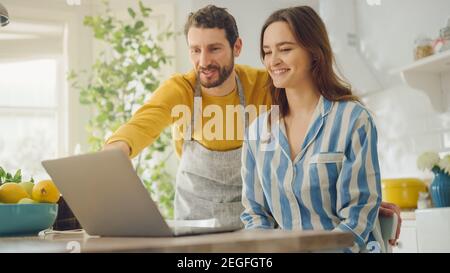 Bella giovane coppia che usa il computer portatile in cucina a casa. Donna in Pajamas e suo marito in un Apron hanno divertimento mentre navigando in Internet prima Foto Stock