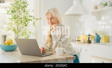Bella donna anziana con computer portatile e bere una tazza di caffè o tè in una cucina soleggiato. Nonna è collegata con i bambini via Foto Stock