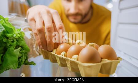 Shot dall'interno cucina Frigo: Bell'uomo apre Frigo porta, guarda all'interno prende poche uova da uova scatola. Uomo che prepara un pasto sano. Punto di vista Foto Stock