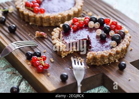 Piccole crostate vegane a base di noci e confettura di bacche decorate con ribes nero, rosso e bianco su chiocciola in legno di quercia Foto Stock