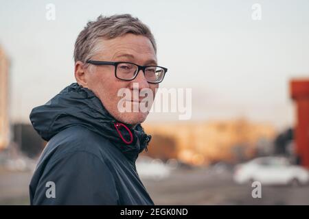 Ritratto di un bell'uomo di mezza età che indossa occhiali. Uomo adulto in giacca all'aperto in città. Sfondo sfocato. Spazio di copia. Foto Stock