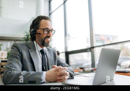 L'operatore di un contact centre felice con cuffie e occhiali utilizza un computer portatile, seduto alla scrivania. Gioioso senior manager sta avendo online business meeting, concetto di comunicazione Foto Stock