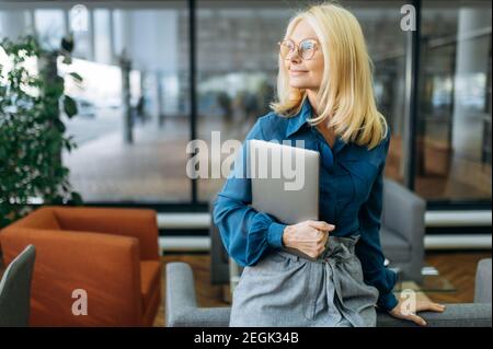 La donna intelligente e sicura si trova in un ufficio moderno, guarda accanto, pensando al progetto di lavoro. Una donna di successo in abbigliamento elegante sta tenendo il computer portatile in braccio Foto Stock