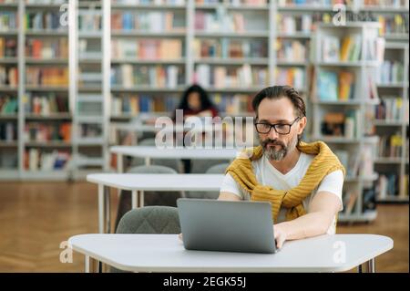 Il freelance maschile concentrato è seduto alla scrivania, utilizza il laptop, sviluppando un nuovo projet. L'uomo moderno anziano sta lavorando al nuovo programma di affari per l'azienda, alla ricerca delle idee Foto Stock
