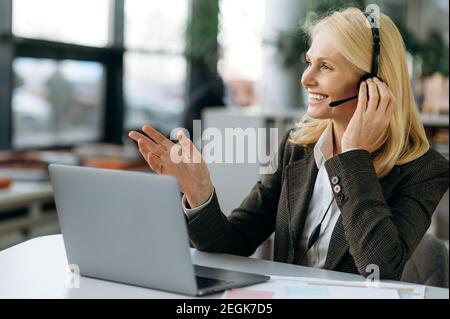 L'operatore femminile amichevole del call center in cuffia comunica con i clienti, consultando le persone. Una donna anziana si siede alla scrivania, usando il computer, facendo una conversazione Foto Stock