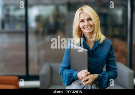 Ritratto amichevole elegante donna di successo sta guardando la fotocamera, tenendo il computer portatile in braccio. Donna d'affari di media età in stile si alza in un ufficio moderno e sorride Foto Stock