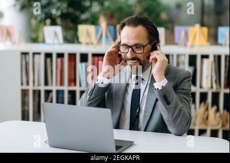 Sorridente operatore maschile di call center in cuffie comunicare con i clienti, consultando le persone. Un uomo d'affari senior seduto alla scrivania, utilizza computr, per conversare Foto Stock