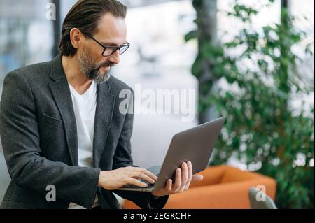 Un dipendente maschile concentrato sta lavorando al progetto, utilizzando il laptop. L'uomo d'affari serio di mezza età è messo a fuoco sul lavoro, cercando alcune idee per l'avvio, la navigazione del Internet Foto Stock