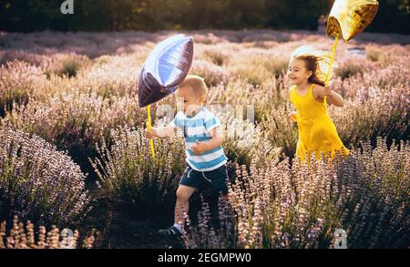 Ragazzo caucasico e sua sorella che corrono in un campo di lavanda tenere palloncini stelle in una giornata di sole Foto Stock