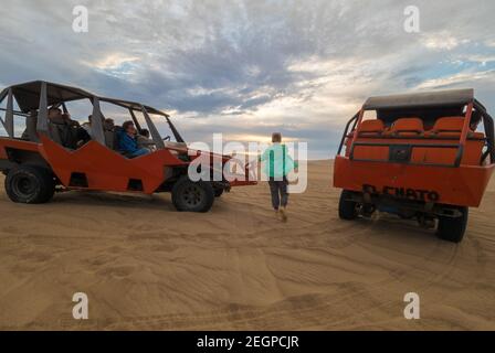 Perù, Ica - 21 settembre 2019 - Donna cammina tra buggy per godersi la vista del tramonto nel deserto, più persone aspettano all'interno delle auto Foto Stock