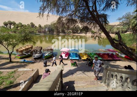 Perù, Huacachina - 1 ottobre 2019 - Oasis lago tour barche colorate ancorate a riva, persone a piedi e dune di sabbia sullo sfondo Foto Stock