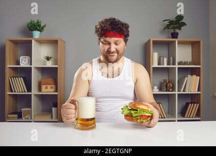 Felice giovane uomo seduto al tavolo, mangiando deliziosi cheeseburger e bevendo birra Foto Stock