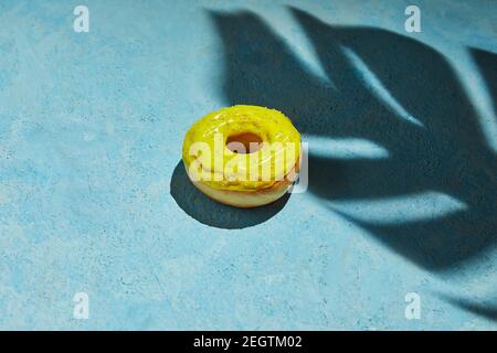 Ciambella con glassa gialla e spruzzi con ombra da foglia di monstera su sfondo blu. Foto Stock