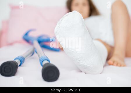 Una ragazza adolescente si trova su un letto con gesso sulla gamba, con una frattura dell'articolazione della caviglia e con stampelle. Gambe fratturate in un bambino.primo piano Foto Stock