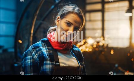 Giovane bella Empowerering Donna con l'orecchio Piercing sorride delicatamente alla macchina fotografica. Autentico Fabricator che indossa abiti da lavoro in un laboratorio di metallo. Scintille Foto Stock