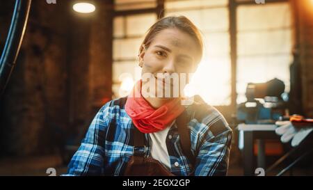 Giovane bella Empowerering Donna con l'orecchio Piercing sorride delicatamente alla macchina fotografica. Autentico Fabricator che indossa abiti da lavoro in un laboratorio di metallo. Foto Stock